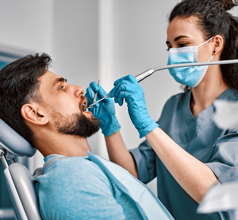 Dentist wearing a mask and gloves examines a man's teeth with dental tools in a clinic setting.