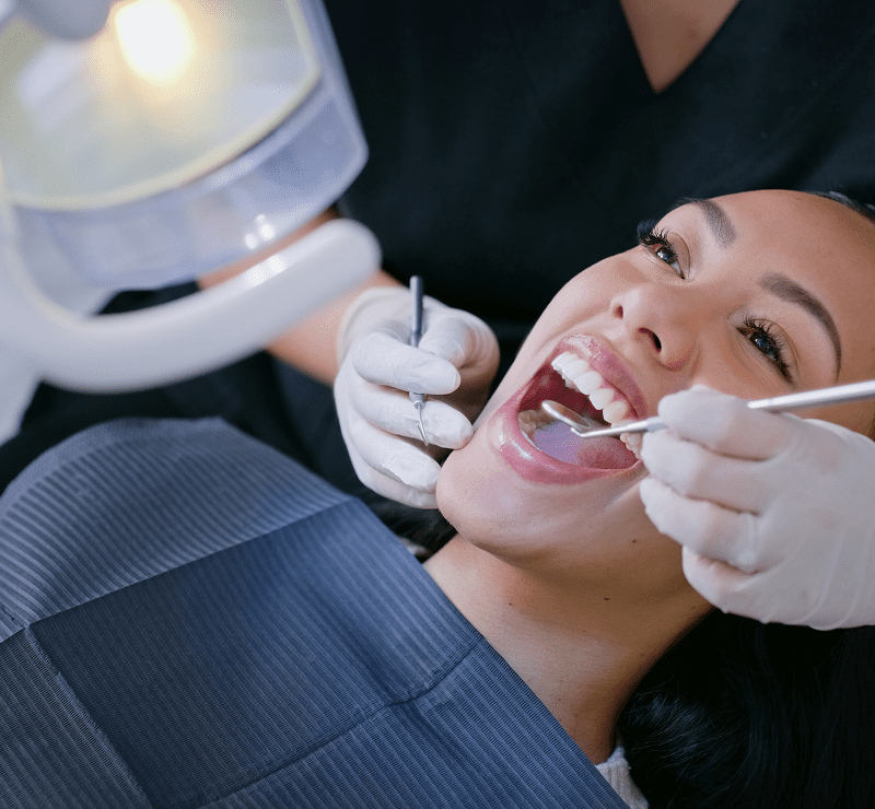 A woman is sitting in a dentist's chair, smiling with her mouth open as a dentist examines her teeth with dental tools under a bright light.