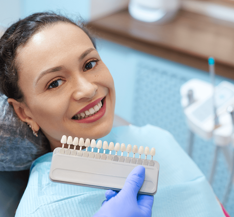 A woman in a dental chair smiles as she looks at the camera. A dentist holds a shade guide against her teeth to match colors.