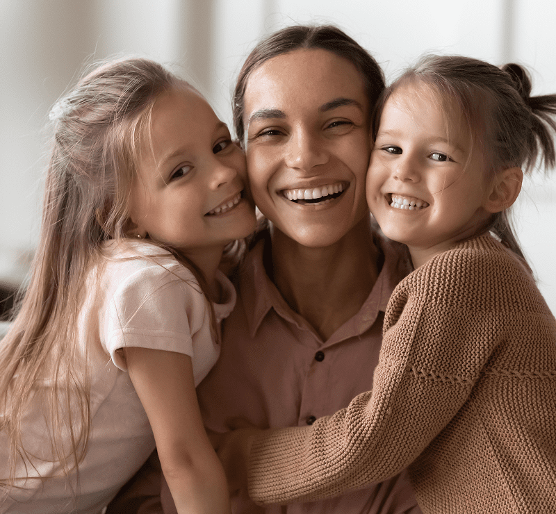 A smiling adult surrounded by two happy children, all embracing and looking at the camera in a warmly lit room.