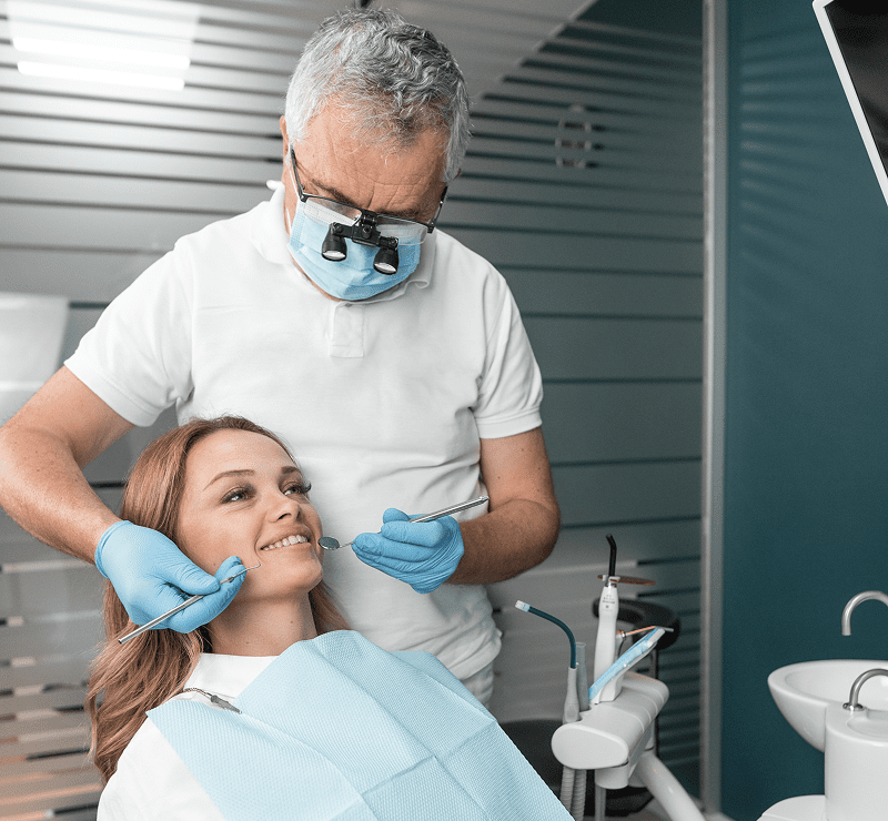 Dentist wearing gloves and magnifying glasses examines a female patient's teeth with dental tools in a clinic.